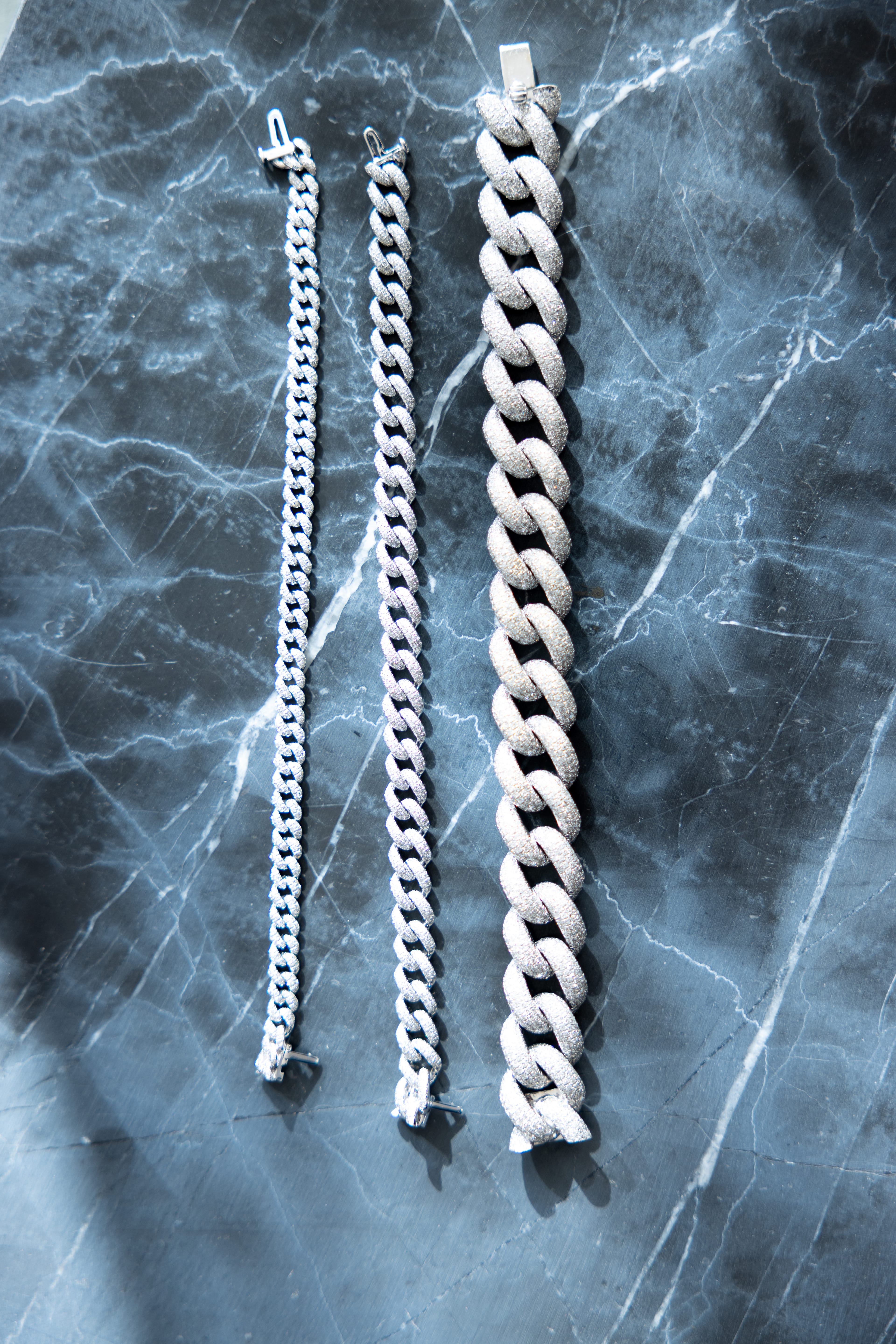 Three silver chain bracelets of varying thicknesses encrusted fully in diamondson a dark, marbled surface.